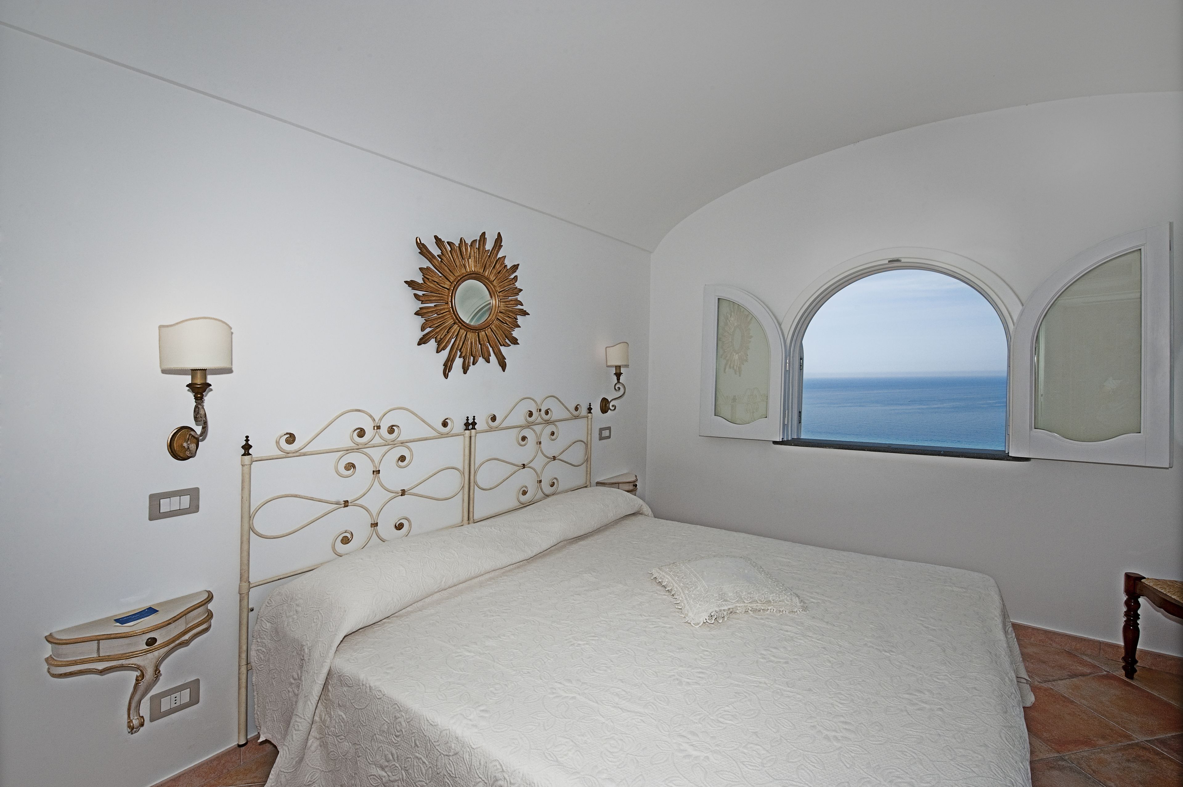 Un dormitorio con cama, tocador con espejo, espejo de sol en la pared y ventana con vista al mar.