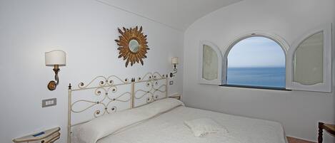 Un dormitorio con cama, tocador con espejo, espejo de sol en la pared y ventana con vista al mar.