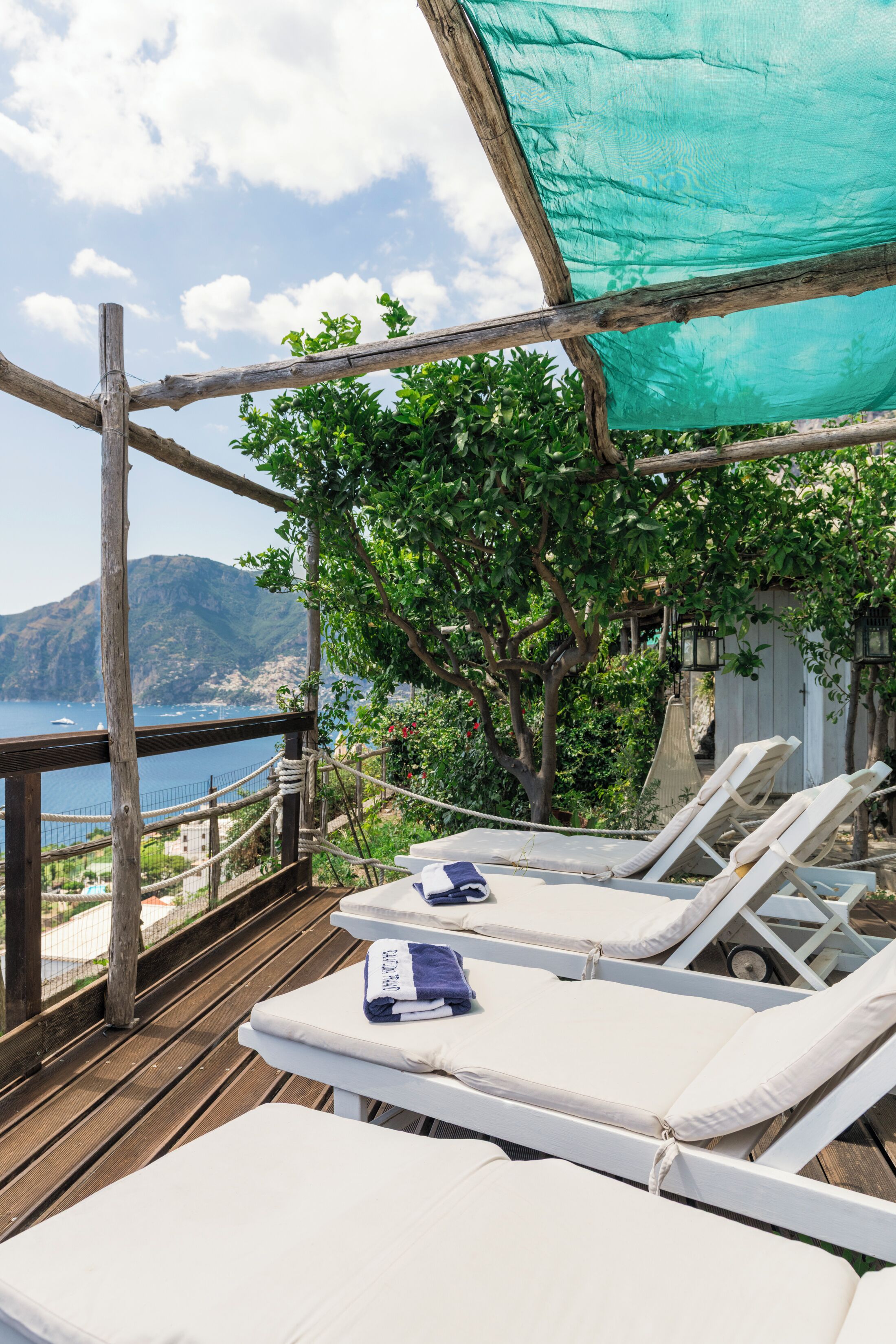 A wooden deck with white lounge chairs and a table, overlooking a coastal area with a view of the sea and mountains.