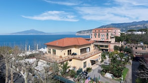 A coastal building with a balcony overlooking the sea, a mountain in the background, and a flag in front. - Al Centro Suites (Sorrento)