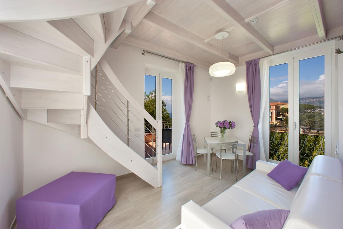 A modern interior with a staircase, a dining area, and a sofa with purple cushions.