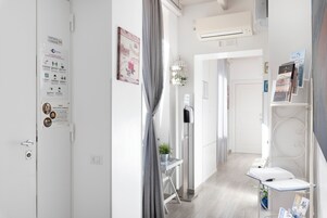 Un pasillo de hotel con una puerta blanca, un tablón de anuncios, una pequeña mesa con una planta y un soporte metálico con folletos.