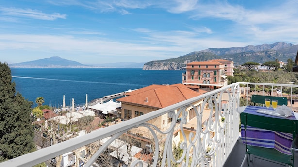 A balcony with a view of the sea and mountains, a building with a red roof, and a table with two glasses of water. - Al Centro Suites (Sorrento)