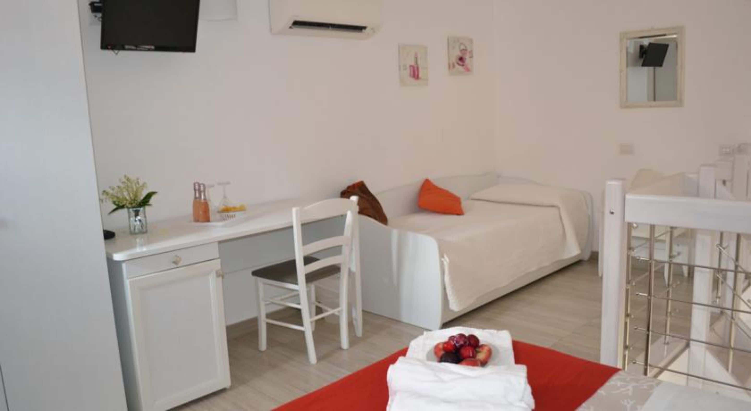 Une pièce moderne et bien éclairée, avec un bureau blanc, une chaise, un lit agrémenté d’un coussin orange et une petite table sur laquelle se trouve un panier de fruits.
