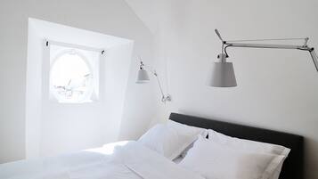 Un dormitorio con cama, ropa de cama blanca, una ventana con vistas y una lámpara de escritorio.