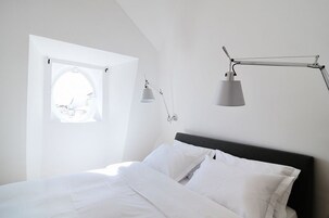 Un dormitorio con cama, ropa de cama blanca, una ventana con vistas y una lámpara de escritorio.