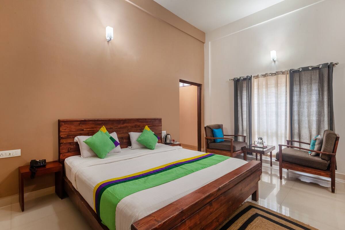Habitación de hotel con una cama de madera, una mesita de noche, una silla, una pequeña mesa y una ventana con cortinas.