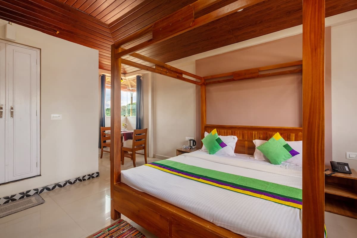 Una cama con dosel y cabecera de madera, una mesita de noche con un teléfono, y una silla en una habitación con piso de baldosas y una puerta al fondo.