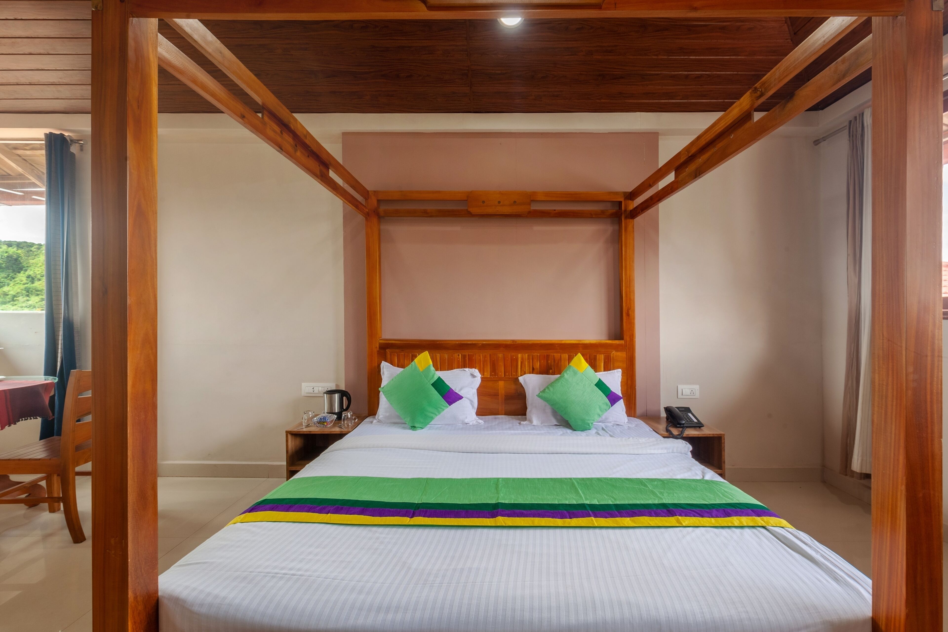 A room with a wooden ceiling, a bed with a striped bedspread, two nightstands, and a view of greenery outside.