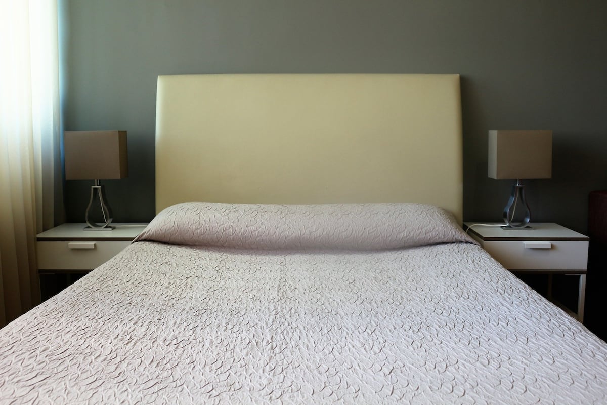 Una cama bien hecha con una colcha blanca texturizada, dos mesitas de noche con lámparas y una pared lisa en una habitación de hotel.
