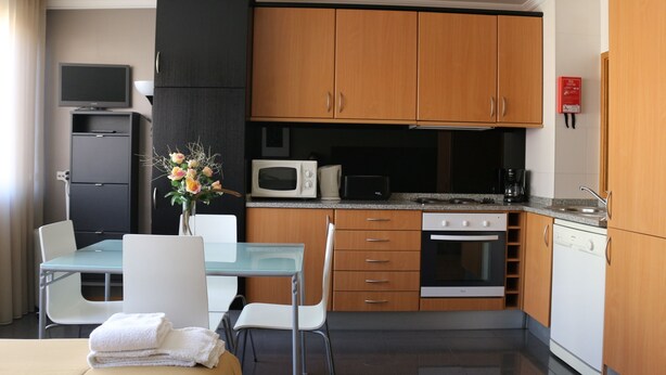 A compact kitchen with wooden cabinets, a microwave, a stove, and a dishwasher. There is a dining table with chairs and a vase of flowers. - Studios Porto Solar (Porto)