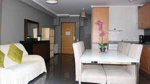 A modern living room with a white sofa, a dining table with chairs, a kitchen area with cabinets, and a hallway leading to a door. - Studios Porto Solar (Porto)
