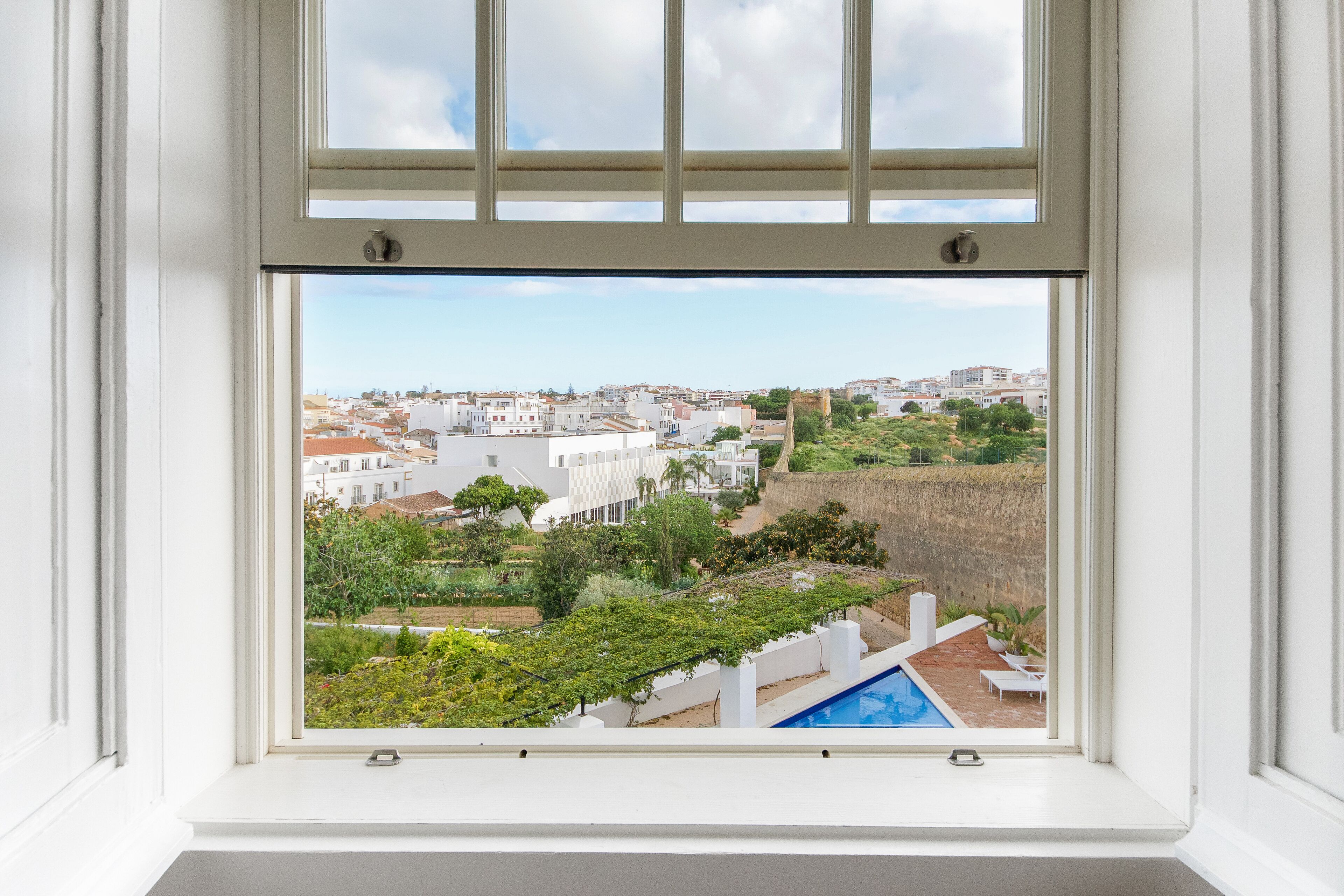 Vista da janela mostrando uma área residencial com piscina, edifícios brancos e vegetação.