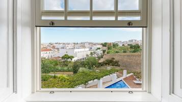 Vista da janela mostrando uma área residencial com piscina, edifícios brancos e vegetação.