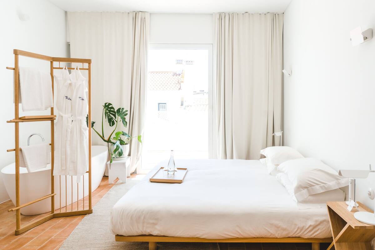 Quarto de hotel com uma cama, mesa de cabeceira de madeira, luminária, banheiro com banheira, varal de madeira e uma planta em vaso.