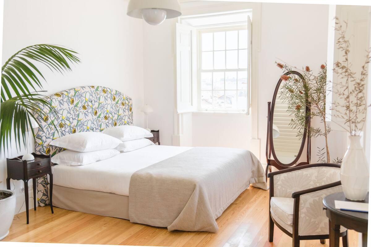 Quarto com cama, mesa de cabeceira, cadeira, espelho, janela e planta em vaso.