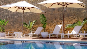 Zona de piscina con sillones reclinables blancos y sombrillas, rodeada por un muro de piedra.