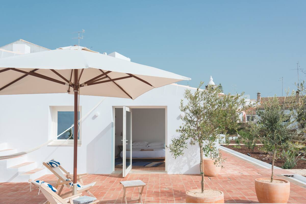 Terraço no telhado com guarda-sol branco, espreguiçadeiras e plantas em vasos.