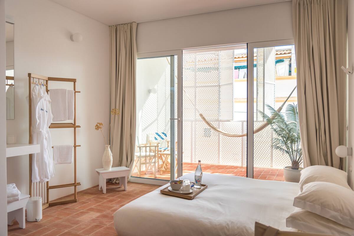 Quarto de hotel com cama, porta-toalhas de madeira, uma pequena mesa com bandeja, planta em vaso e vista para a área externa por uma porta de vidro deslizante.