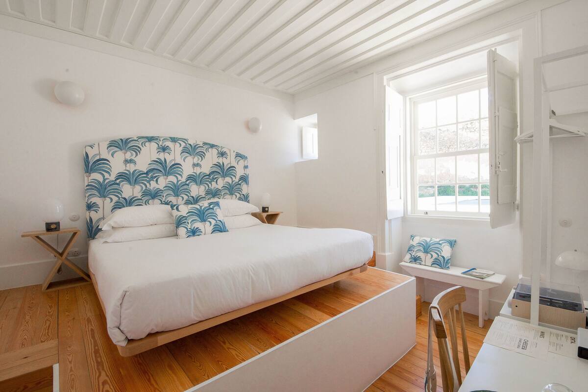 Quarto com cama, piso de madeira, paredes brancas, uma janela e uma escrivaninha com cadeira.