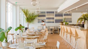 Un restaurante moderno con mesas y sillas de madera, una zona de bar con taburetes y amplios ventanales que permiten la entrada de luz natural.