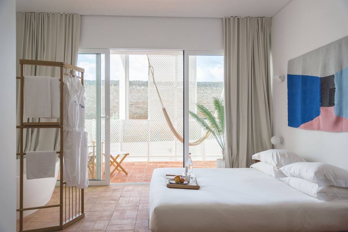 Quarto de hotel com cama, porta-toalhas, bandeja com frutas e vista para o exterior.