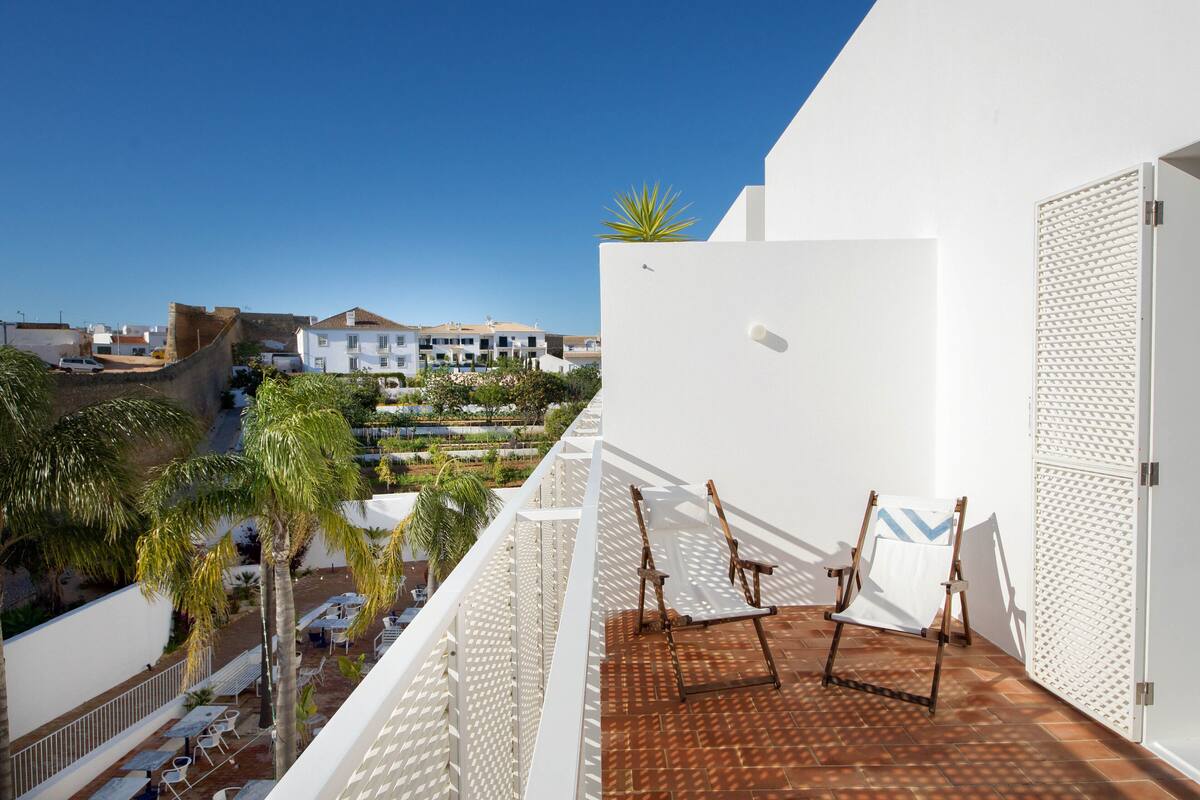 Varanda com guarda-corpo branco, uma cadeira de madeira e vista para área residencial com casas e palmeiras.