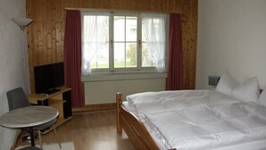 A room with a wooden ceiling, a bed with white bedding, a television on a wooden stand, a small round table, and a chair.