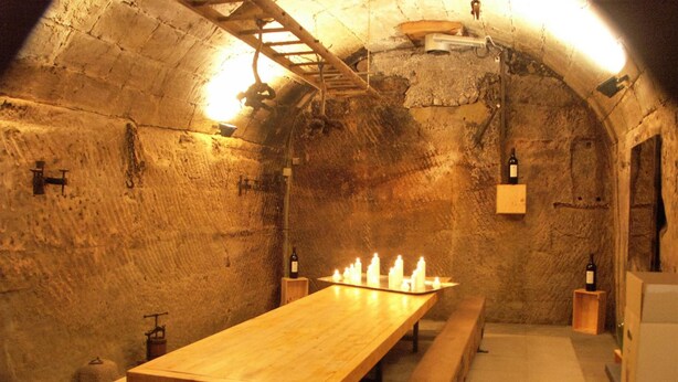 A rustic underground room with a long wooden table and benches, stone walls, and hanging light fixtures.