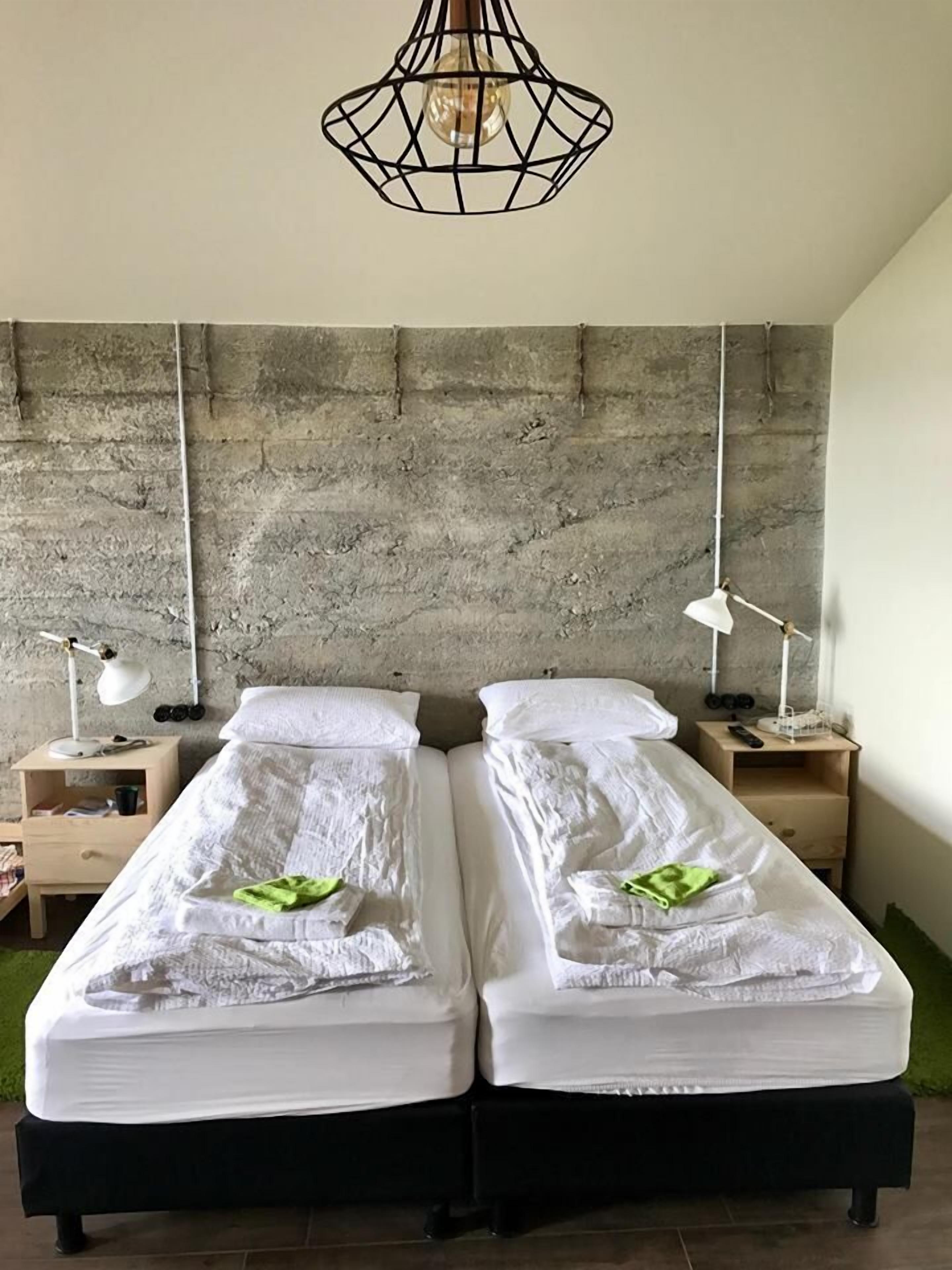 Habitación con dos camas, pared de concreto, lámpara colgante y mesitas de noche con lámparas.