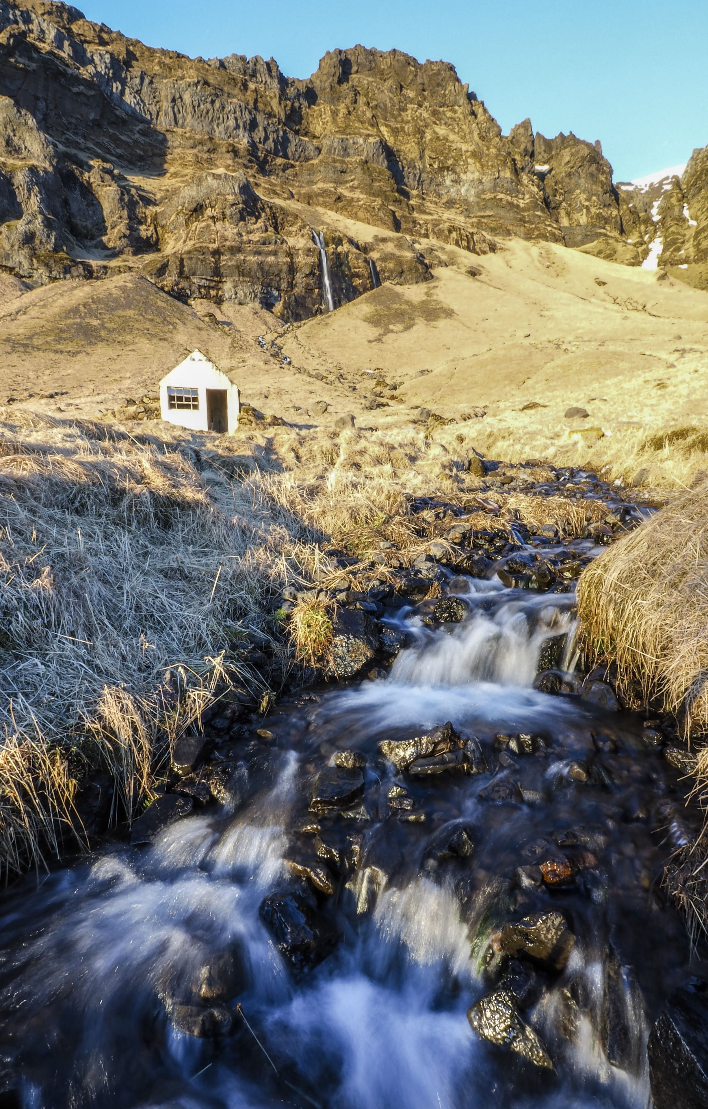 A small white building with a window, a flowing stream, and rocky terrain.