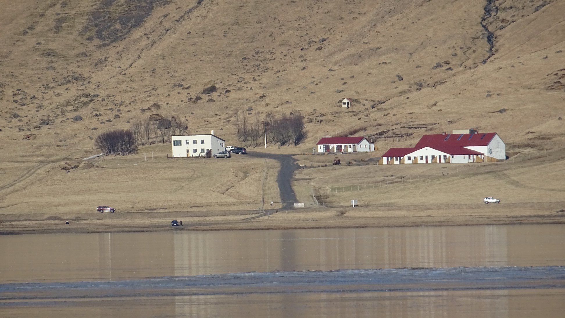 Un paysage rural avec une maison blanche et un bâtiment à toit rouge, un chemin de terre et une étendue d’eau.