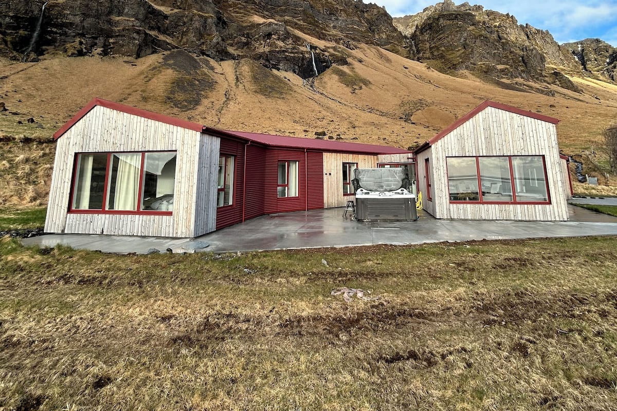 A wooden cabin with a red roof and large windows, situated in a mountainous area with a clear sky.