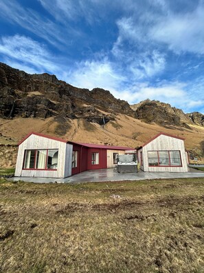 A wooden cabin with a red roof and large windows, situated in a mountainous area with a clear sky. - The Garage (Rangárþing eystra)