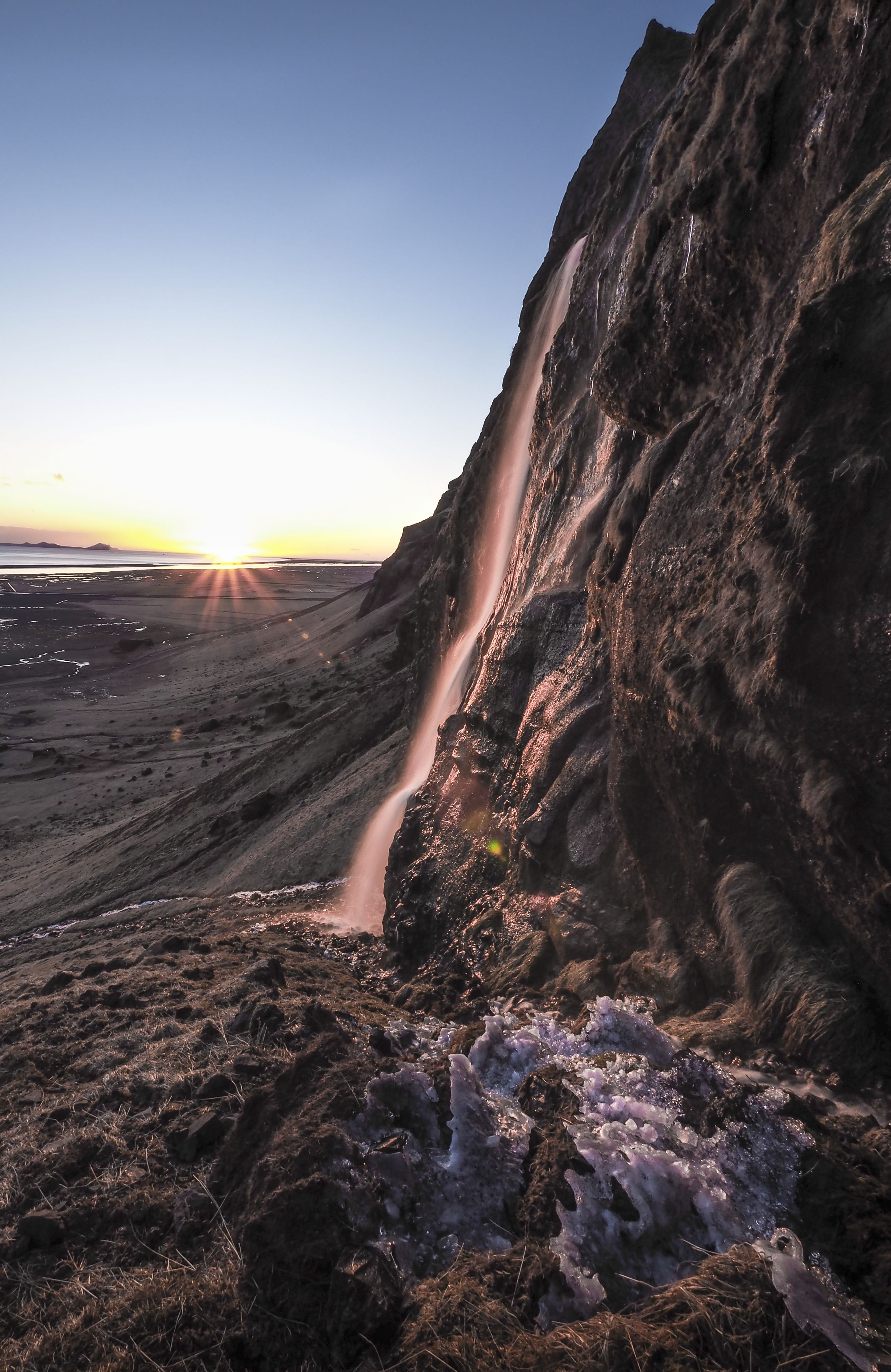 Um penhasco acidentado com uma cascata descendo sua lateral, rodeado por terreno rochoso e vegetação escassa.
