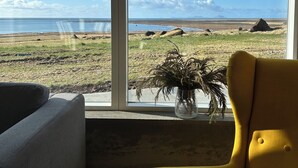 A room with a large window offering a view of a beach and distant mountains. - The Garage (Rangárþing eystra)