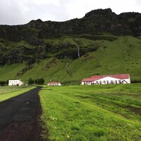 Paisaje rural con una carretera que lleva a un edificio blanco de techo rojo, rodeado de campos verdes y una montaña al fondo.
