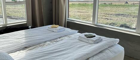 A bedroom with a large bed, two windows with curtains, and a view of the beach and sea.