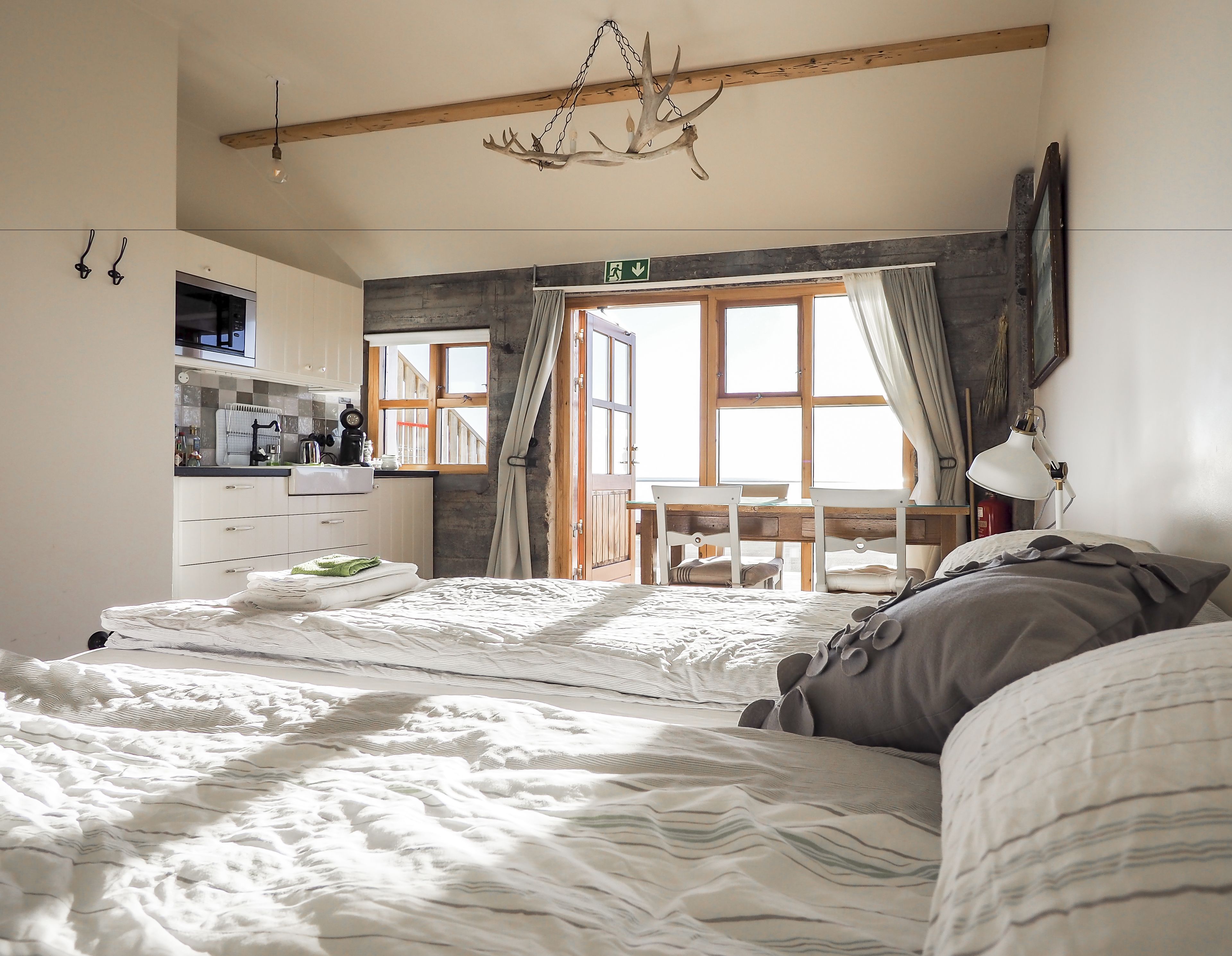 A bedroom with a large bed, a desk, and a chair. There is a window with curtains, a chandelier, and a view of the outside.