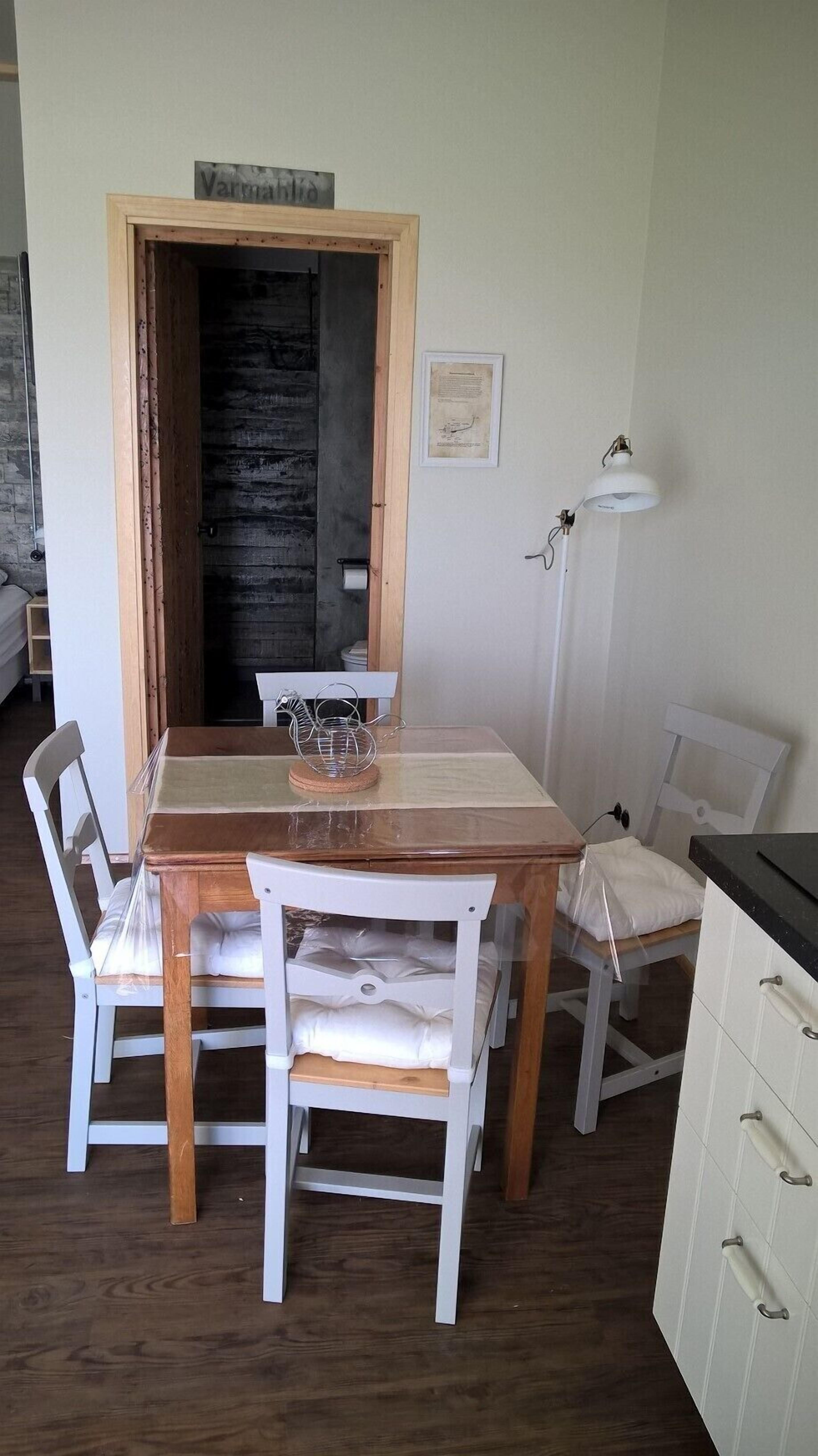 A dining area with a wooden table and white chairs, a small kitchen area with cabinets, and a door leading to another room.