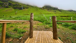 A wooden deck with a view of a green hillside and a misty mountain in the background. - The Garage (Rangárþing eystra)