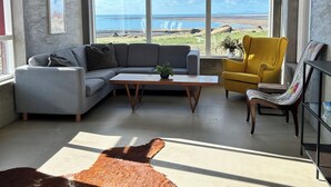 A modern living room with a gray sofa, a yellow armchair, and a wooden coffee table. There is a large window offering a view of the beach and ocean. - The Garage (Rangárþing eystra)
