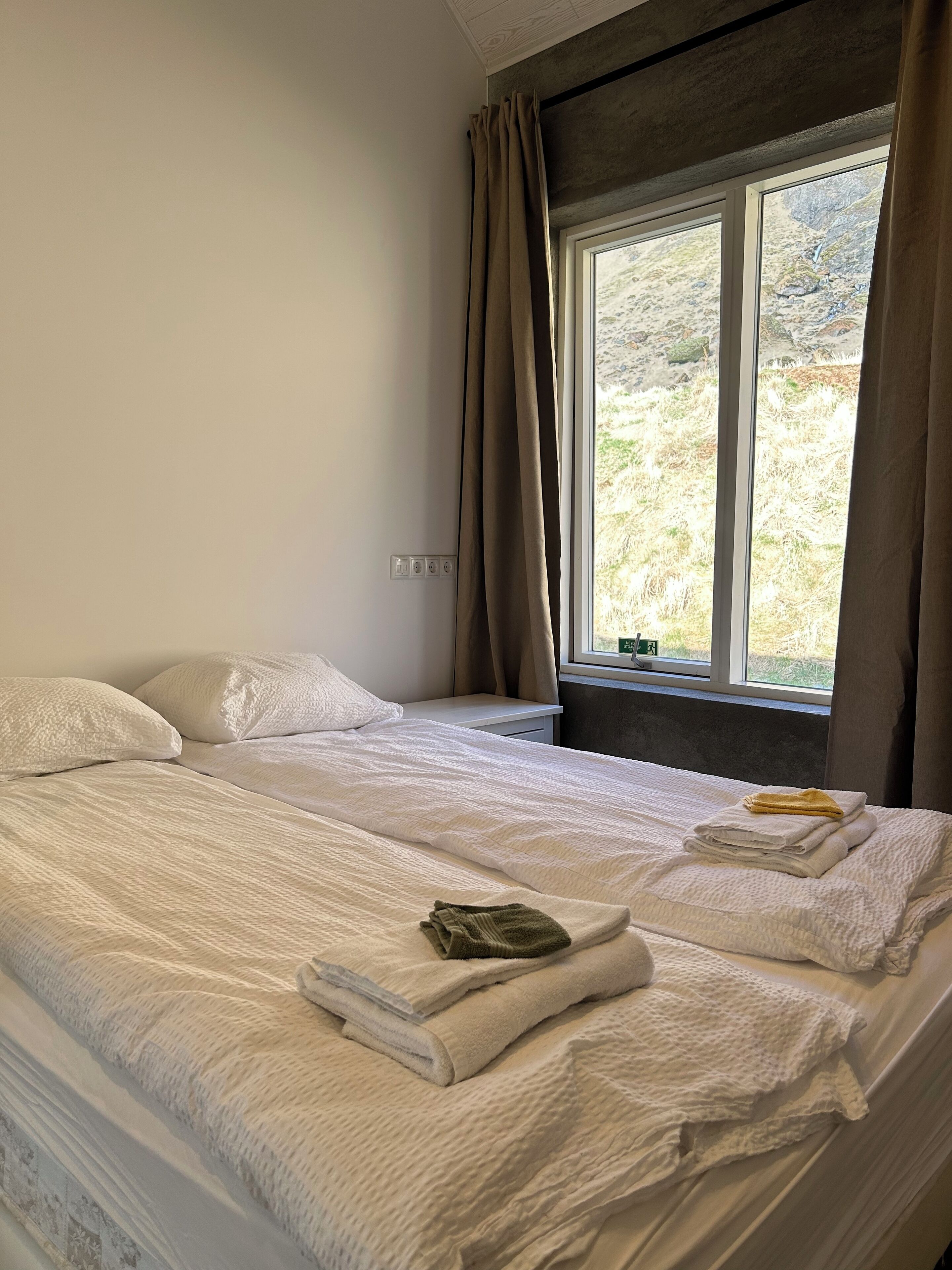 A bedroom with two beds, a window with curtains, and a view of a mountainous landscape.