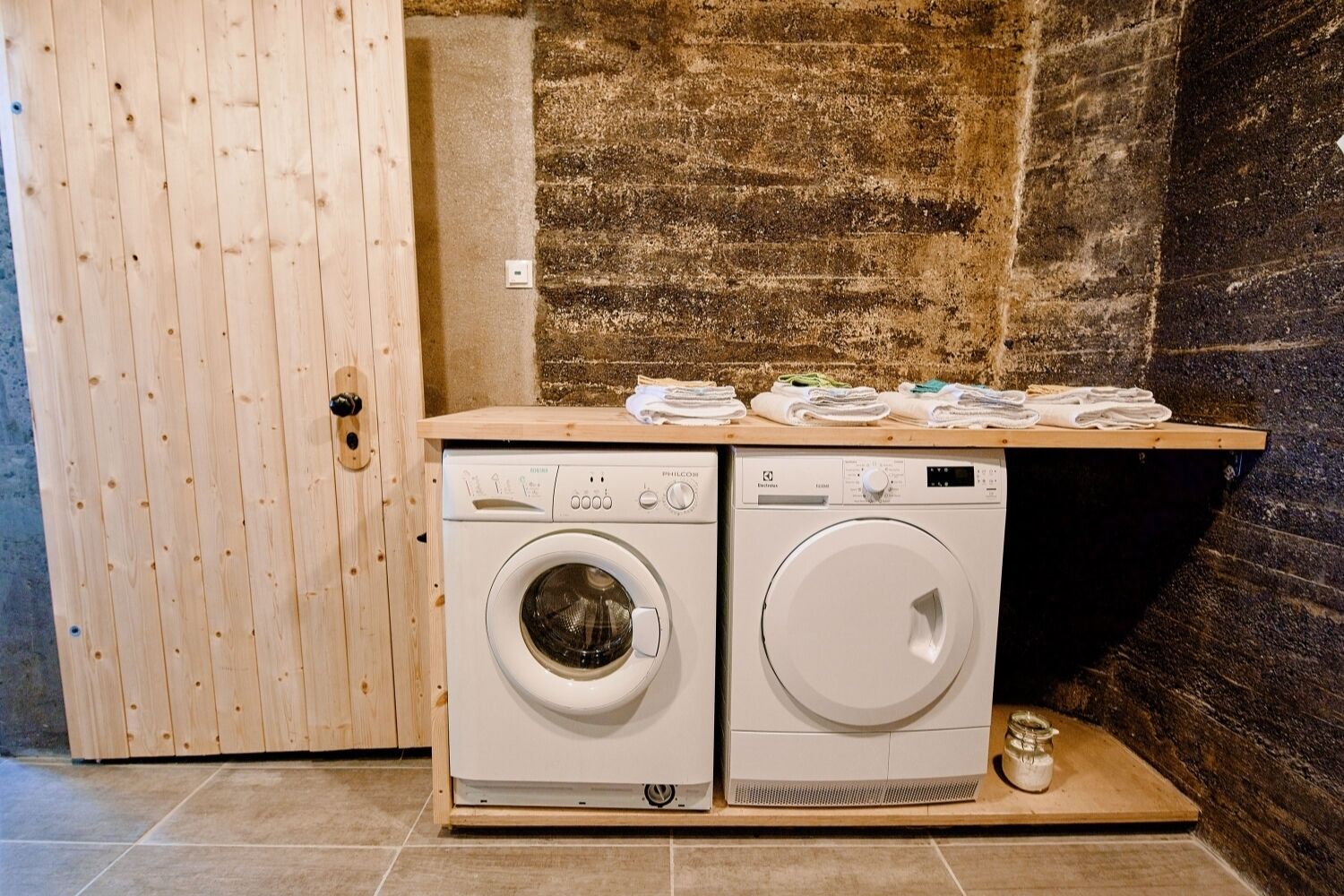 A laundry area with two front-loading washing machines and a wooden shelf above them.