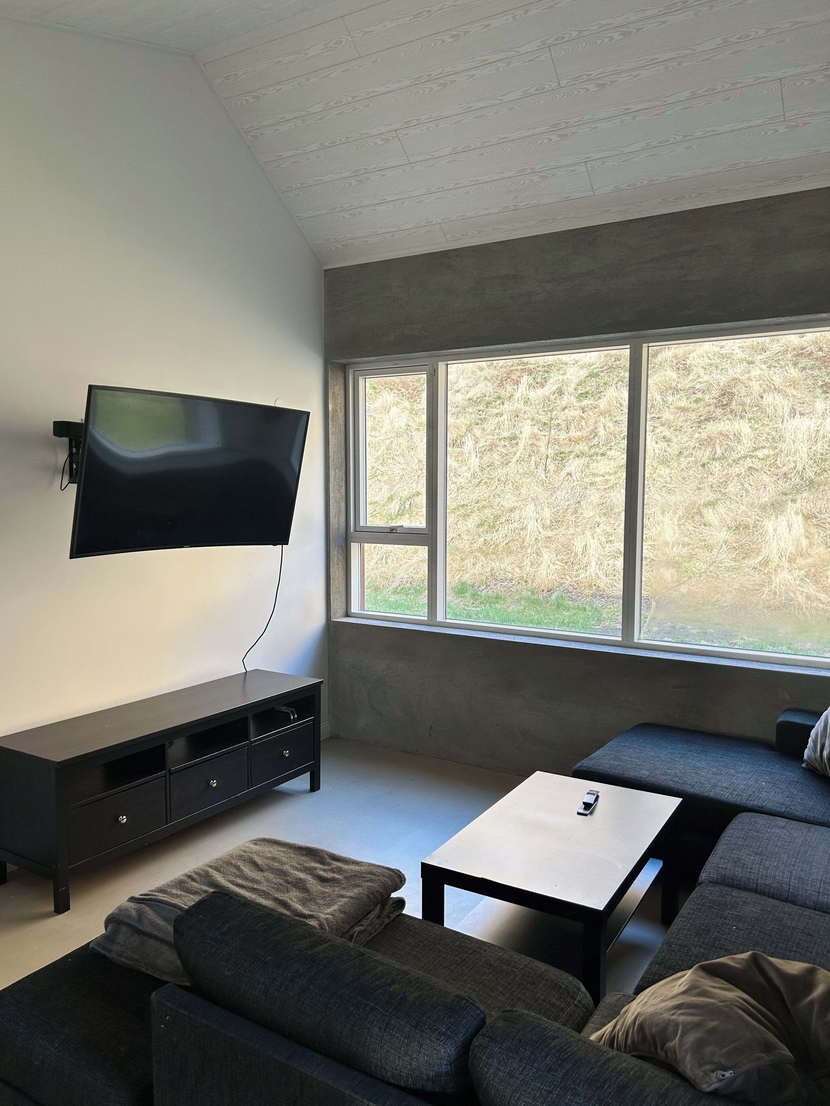 A modern living room with a flat-screen TV mounted on the wall, a dark wooden entertainment unit, a grey sofa, a small table, and a large window offering a view of a grassy area.