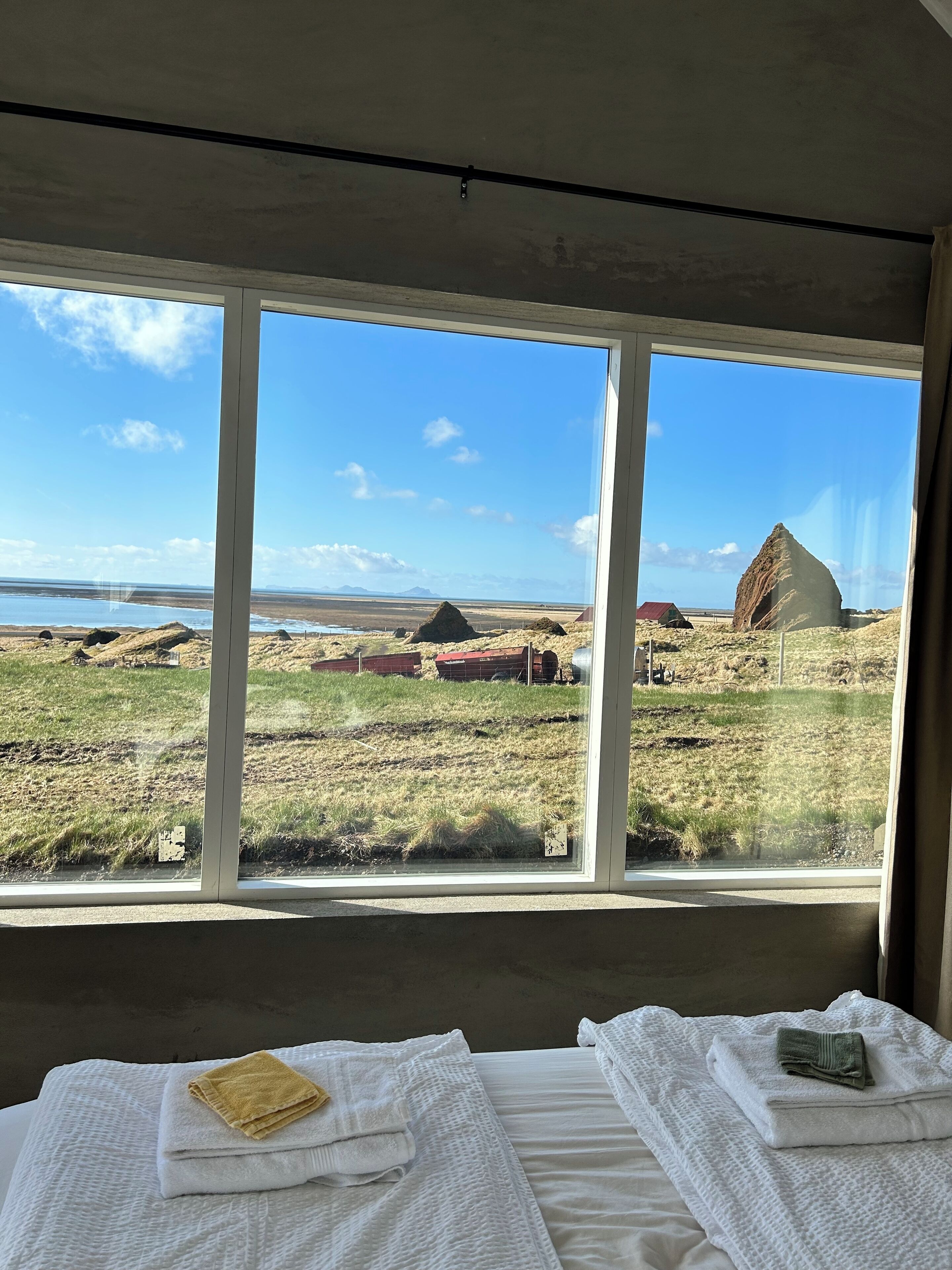 A room with a large window offering a view of a grassy landscape with a distinctive rock formation and a red building in the distance.