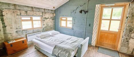 Un dormitorio con piso de madera, una cama con ropa de cama blanca, un aparador de madera y una puerta con ventana.