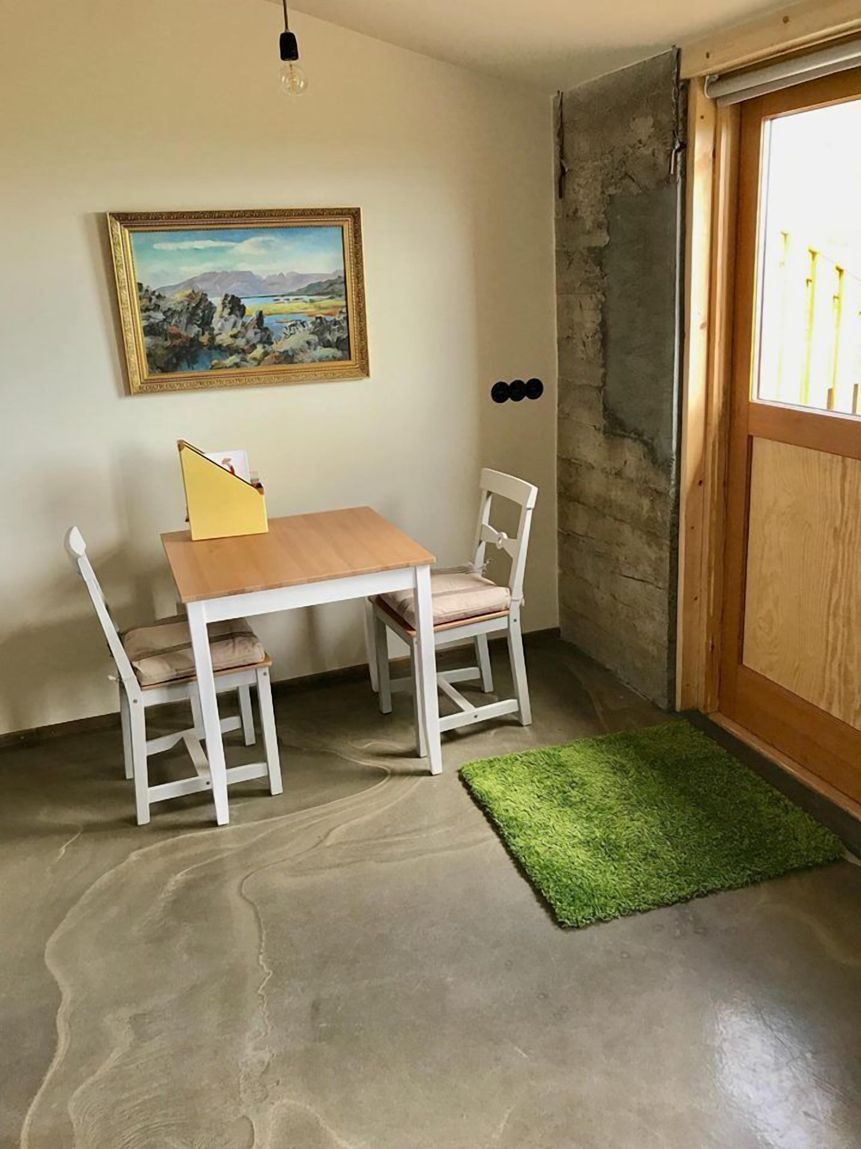 Et værelse med et træbord og to hvide stole, en grøn måtte, et billede på væggen og en trædør.