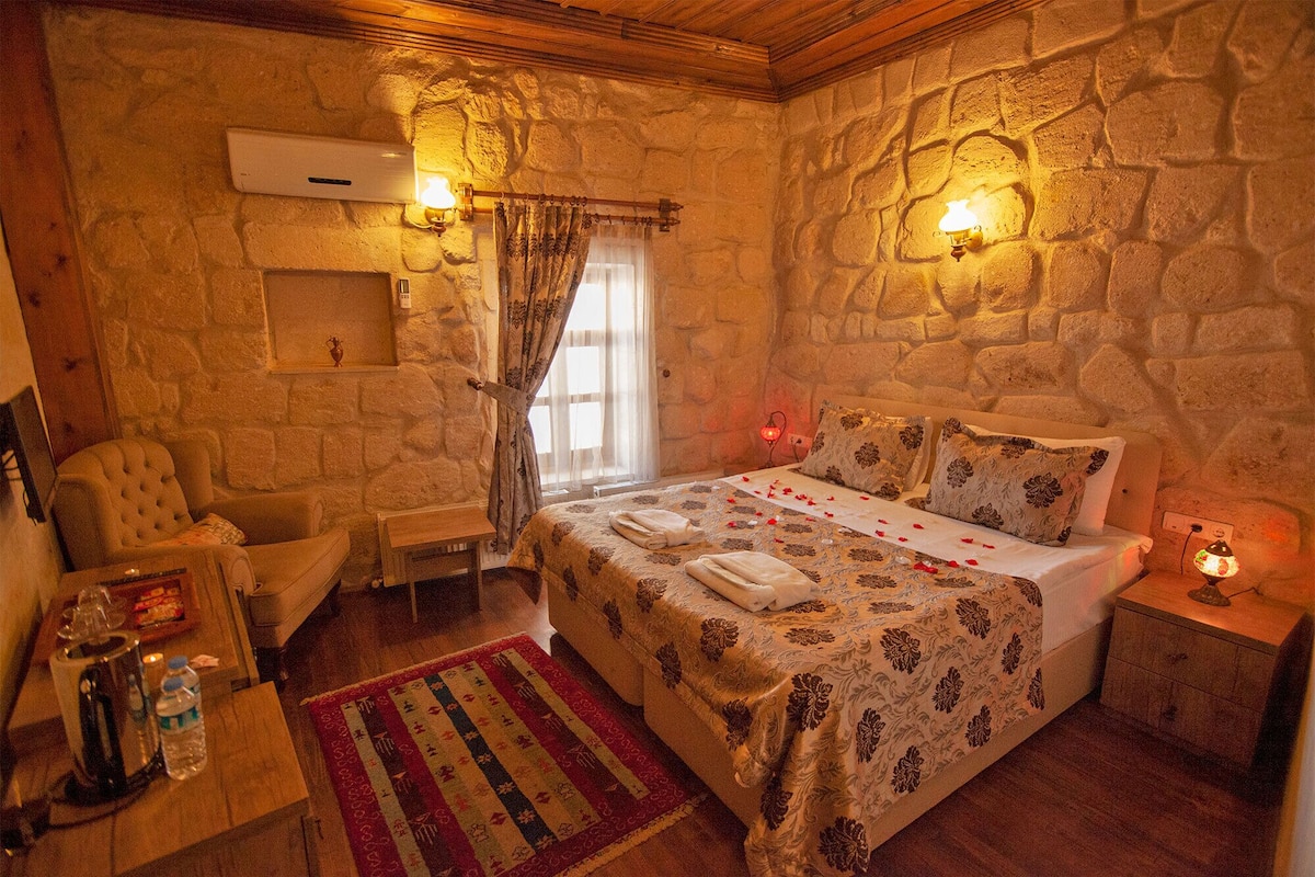 A cozy bedroom with a stone wall, a bed with floral bedding, a wooden chair, a small table with a kettle and water bottle, and a colorful rug.