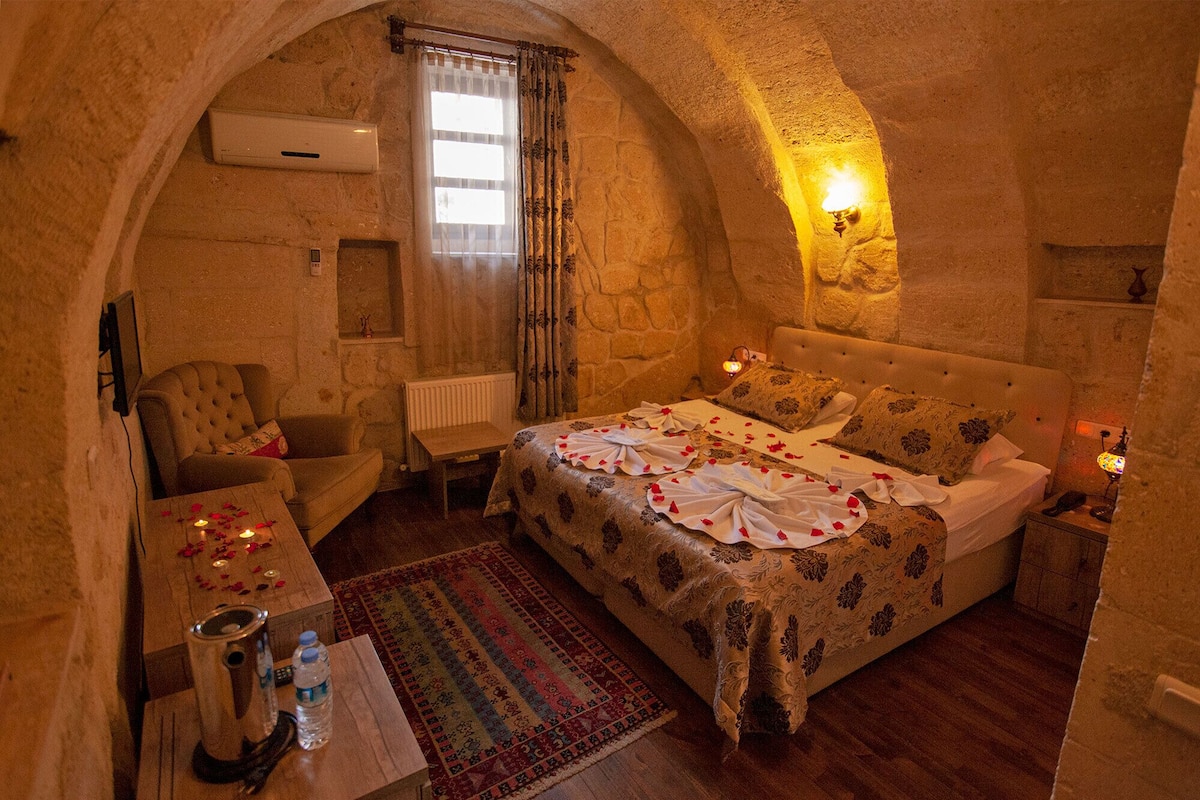 A cozy bedroom with a stone wall, a bed with rose petals, a small table with a water bottle, and a chair.